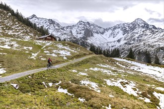 Two mountain bikers riding on path past wooden cabin on mountain slope with patches of melted snow in Montafon region in autumn