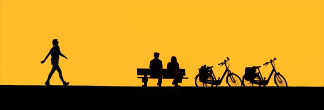 Girl walking past couple of elderly cyclists resting on bench next to their two bicycles silhouetted against yellow sunset sky in summer