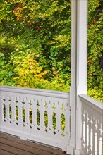 Railing on a porch with white wooden carpentry by a deciduous trees in autumn colors