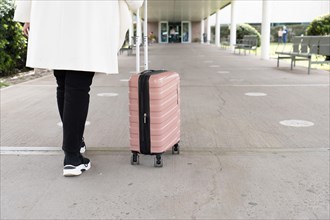 Shot of legs of woman in sneakers and coat walking to main entrance of airport and rolling her wheeled suitcase. Travel concept