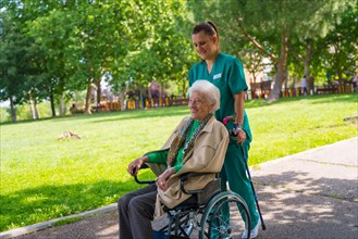 An elderly woman with the nurse on a walk through the garden of a nursing home in a wheelchair next to nature and trees