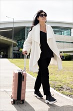 Full shot of business woman with sunglasses leaving the airport carrying her luggage. Concept of business travel