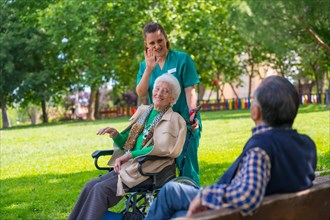 An elderly woman with the nurse on a walk through the garden of a nursing home in a wheelchair and greeting an elderly man