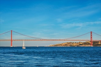 View of 25 de Abril Bridge famous tourist landmark over Tagus river and a tourist yacht boat at sunset. Lisbon