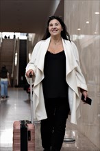 Elegant mature woman walking and pulling her suitcase in airport terminal. Portrait of business woman smiling and looking at camera carrying her wheeled suitcase at airport