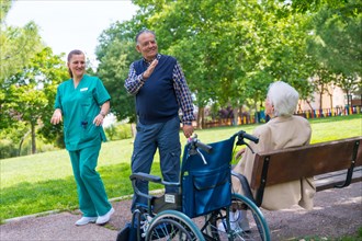 An elderly man with the nurse on a walk through the garden of a nursing home greeting an elderly woman in a wheelchair