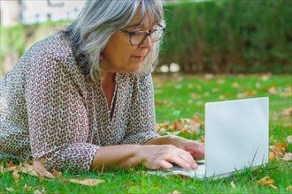 Woman with white hair and glasses lying on the grass in a park with a laptop computer