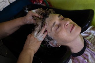 Close-up of mature client having her hair washed by hairdresser after coloring her hair in beauty salon. Small Latin beauty salon business