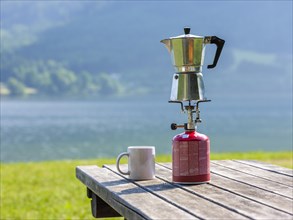 Espresso pot on a gas cooker next to a cup standing on a wooden table at the lake