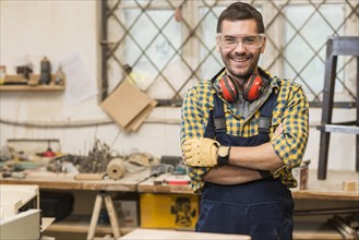 Smiling male carpenter wearing safety glasses standing front workbench his arm crossed