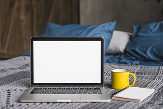 Laptop blank white screen near cup spiral notepad bed
