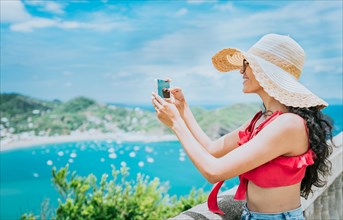 Traveler girl photographing a bay from a viewpoint. Happy tourist woman in a hat photographing a viewpoint