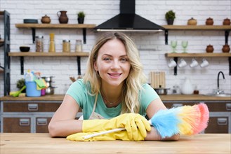 Smiling housekeeper leaning kitchen counter holding feather duster looking camera