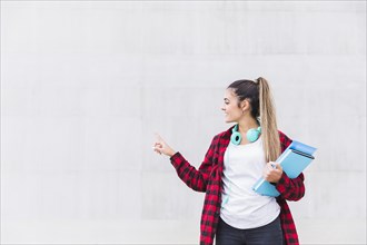 Portrait smiling female university student holding books hand pointing her finger white wall with copy space