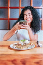 Happy girl photographing a plate of chocolate crepe and ice cream. Young woman using cell phone and taking a picture of chocolate crepe