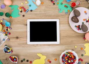 Blank digital tablet surrounded with colorful gems candies easter eggs paper cutout bunny wooden table