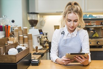 Blonde young woman standing coffee shop counter looking digital tablet