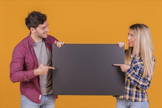 Portrait young couple pointing their finger blank black placard against orange backdrop
