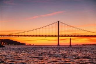 View of 25 de Abril Bridge famous tourist landmark of Lisbon connecting Lisboa and Almada over Tagus river with tourist yacht silhouette at sunset. Lisbon