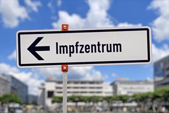 White German street sign pointing towards vaccination center called 'Impfzentrum' set up to vaccine people against Corona virus in front of blurry city background