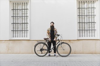 Bearded young man standing with bicycle against wall