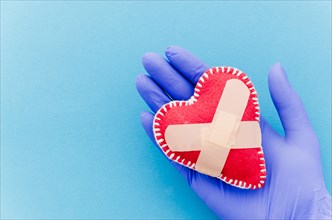 Close up hand wearing surgical gloves holding heart shaped stitched textile heart with crossed bandages blue backdrop