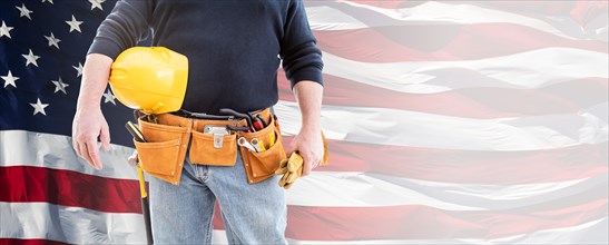 Male contractor wearing tool belt and holding blank yellow hardhat and gloves over waving american flag background banner