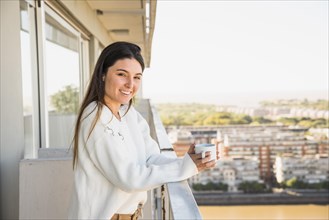 Portrait smiling young woman standing balcony holding white coffee cup