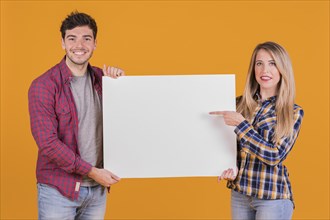 Young woman pointing her finger placard hold by his boyfriend against orange background