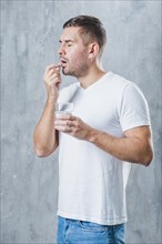 Sick young man standing against gray backdrop holding water glass hand taking pill