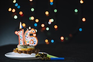 Number 16 birthday lighted candle slice tart with party horn blower against illuminated bokeh backdrop