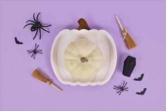 Cream colored pumpkin on plate surrounded by Halloween decoration on violet background