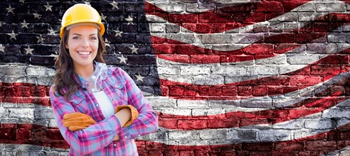 Female contractor wearing blank yellow hardhat and gloves over waving american flag on brick wall background banner