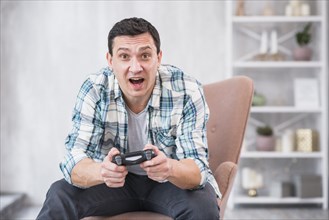 Excited young male sitting armchair playing with gamepad