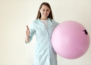 Smiley physiotherapist holding exercise ball giving thumbs up