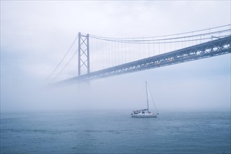 View of 25 de Abril Bridge famous tourist landmark of Lisbon connecting Lisboa and Almada in heavy fog mist wtih yacht boats passing under. Lisbon