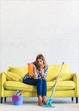 Sad young woman sitting yellow sofa looking orange rubber gloves