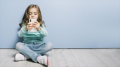 Portrait little girl sitting hardwood floor looking smartphone