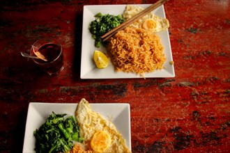 Stir fried wonton noodles with fried egg and spinach against a rustic red table