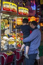 Father with son looking at claw crane game machine filled with toys in amusement arcade at travelling funfair
