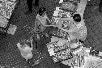 Black and white bird's eye view photo of a stall full of fish with the seller and a buyer