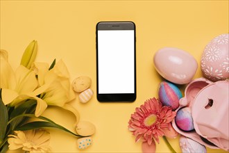 White blank screen mobile phone decorated with lily gerbera flower colorful easter eggs yellow background