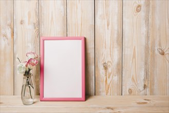 Flower vase near white blank frame with pink border against wooden wall