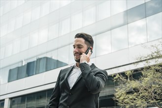 Smiling young businessman standing front office building talking mobile phone