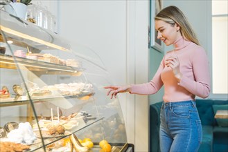 Smiling young woman choosing cake standing front glass cabinet