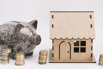Ceramic piggybank with stack coins near cardboard house white background