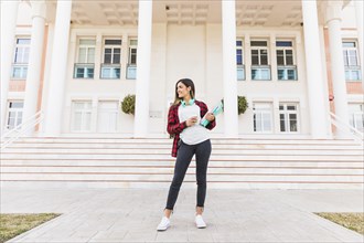 Portrait smiling teenage female student holding books takeaway coffee cup standing front university building