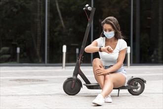 Woman with medical mask checking smartwatch while sitting electric scooter