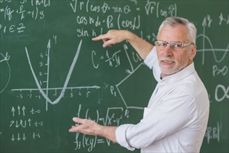 Senior teacher glasses explaining math example green chalkboard