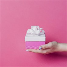 Female s hand holding box with white ribbon bow pink background